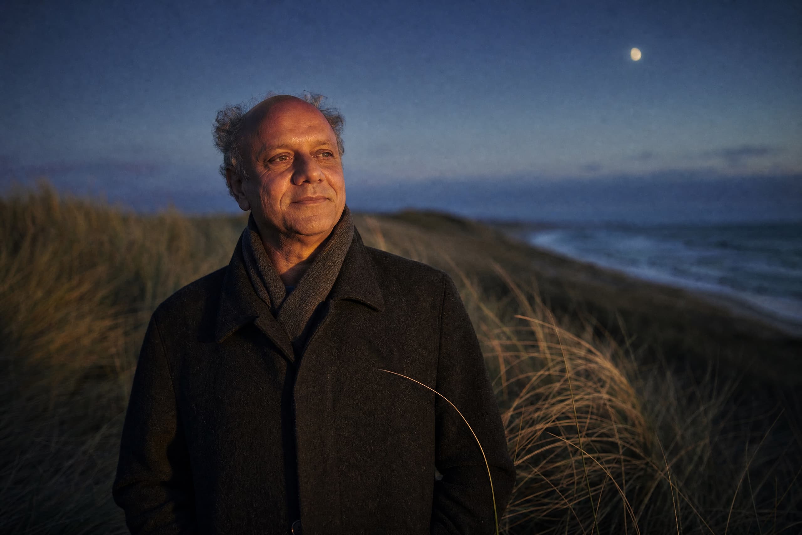 Esam Abou-El Magd steht zur blauen Stunde im Dünengras an der Ostsee und blickt aufs Meer; am Himmel ein kleiner Mond.